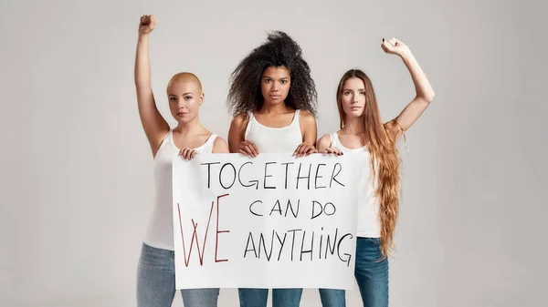 Tres mujeres diversas confiadas en camisa blanca y pantalones vaqueros que parecen serios mientras sostienen, de pie con una pancarta en sus manos aisladas sobre un fondo gris — Foto de Stock