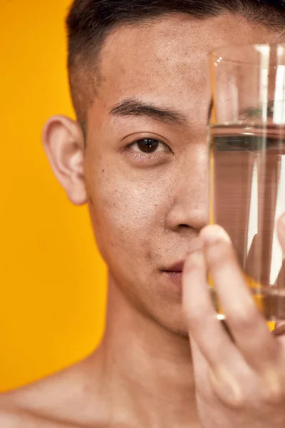 Close-up portret van de jonge aziatische man kijken naar de camera, het houden van een glas water in de buurt van zijn gezicht geïsoleerd over gele achtergrond. Gezondheidszorg, welzijnsconcept — Stockfoto