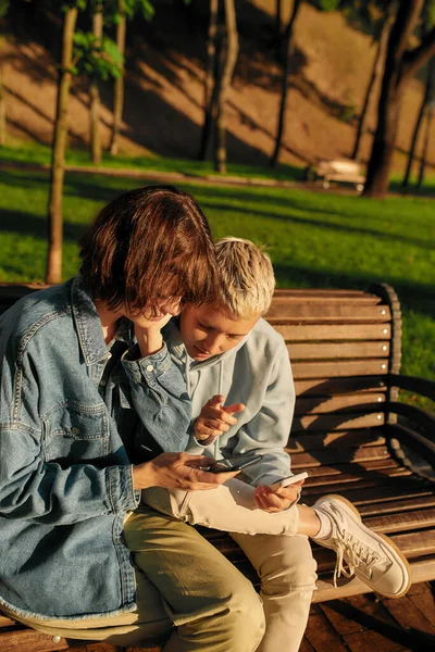Two young women holding their smartphone, sitting on the bench in the park. Lesbian couple using their phones while spending time together outdoors