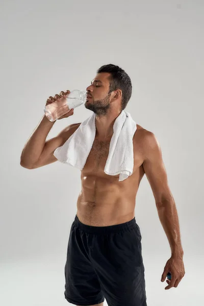 Sentindo sede após o treino. tiro vertical de jovem homem musculoso bonito com toalha em seus ombros beber água enquanto em pé isolado sobre fundo cinza. — Fotografia de Stock