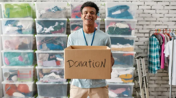 寄付箱を持っている陽気な男とカメラで笑顔,服でいっぱいのラックやボックスの前でポーズ,慈善のために働く若い男性ボランティア — ストック写真