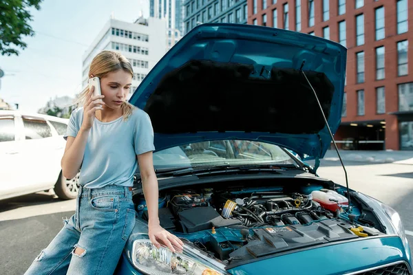 Aantrekkelijke jonge vrouw belt hulpdiensten, praat aan de telefoon terwijl ze staat, leunt op haar kapotte auto met open motorkap op de straat van de stad — Stockfoto