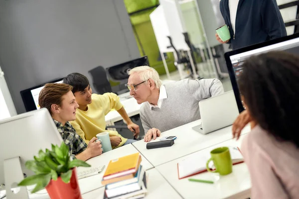 Åldrad man, seniorpraktikant chattar med sina unga kollegor, Vänliga arbetare pratar med nyanställd under fikapausen på jobbet — Stockfoto