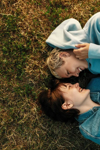 Cropped shot of happy lesbian couple looking at each other and laughing while lying on the grass in summer park. Girls in love cuddling, spending time together outdoors — Stock Photo, Image