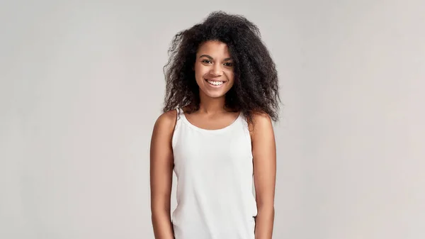 Ritratto di giovane donna afroamericana con i capelli ricci che indossa camicia bianca sorridente alla macchina fotografica mentre in piedi isolato su sfondo grigio — Foto Stock