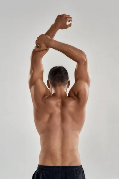 Rear view of muscular man with naked torso raising his arms above head isolated over grey background, posing in studio — Stock Photo, Image