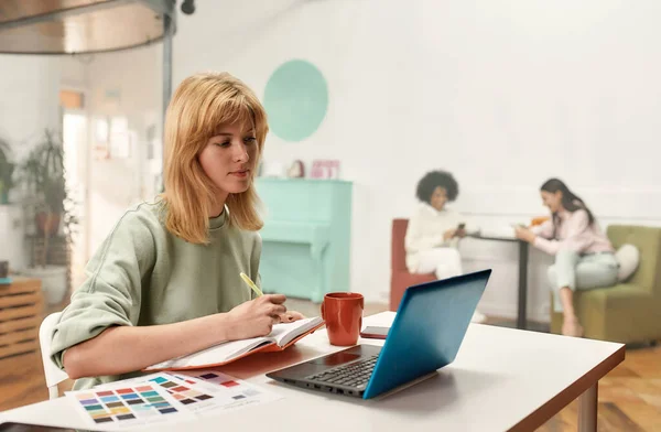 Творческая молодая женщина со светлыми волосами, сидящая и работающая над своим ноутбуком, пока ее коллеги сидят вместе на заднем плане — стоковое фото