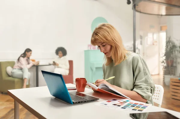 Egy csinos szőke ül egy asztalnál egy laptop előtt, miközben fiatal kollégáit a háttérben dolgoztatja. — Stock Fotó