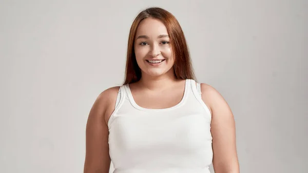 Ritratto di una bella giovane caucasica plus size donna vestita di camicia bianca, sorridente alla macchina fotografica mentre posa isolata su sfondo grigio — Foto Stock