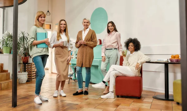 Farklı yaşlarda beş yaratıcı kadından oluşan bir grup sabah toplantısında şık giyinmiş, gülümseyen ve kameraya bakan bir grup.