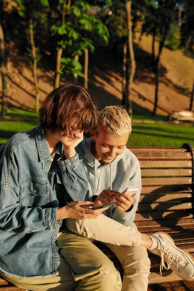 Dos mujeres alegres sosteniendo su smartphone, sentadas en el banco del parque. Pareja lesbiana usando sus teléfonos mientras pasan tiempo juntos al aire libre —  Fotos de Stock