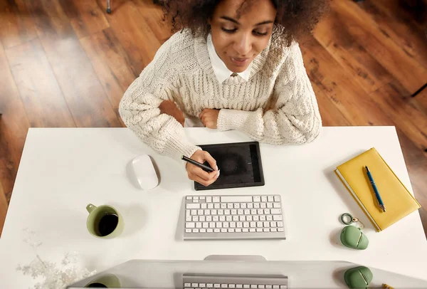 Egy jól öltözött, sötét bőrű hölgy, aki egy grafikus táblával dolgozik egy számítógép előtt, aki egy asztalnál ül. — Stock Fotó