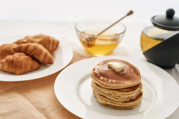 Close up of a stack of sweet tasty pancakes, croissants, honey in a bowl with dipper and a teapot, Breakfast served on the table over white background — Stock Photo, Image