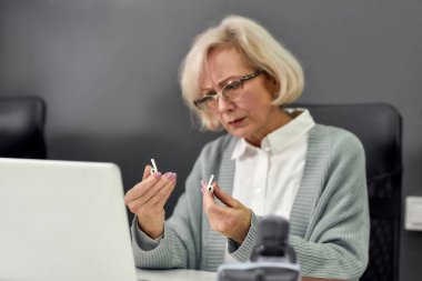 Yaşlanmış kadın portresi, kıdemli stajyer, kablosuz kulaklıklara bakmak, dizüstü bilgisayar kullanmak, masa başında oturmak, modern ofiste çalışmak.