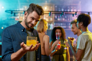 Elinde bir kokteylle kameraya bakan yakışıklı genç adam ve sohbet eden arkadaşları, arka plandaki bar tezgahında içki içiyorlar.