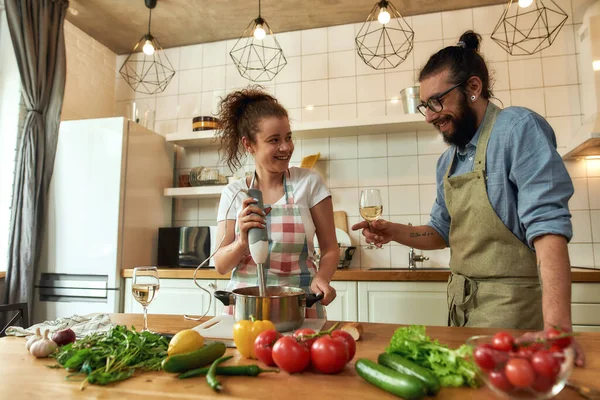 Ιταλός, σεφ μαγειρεύουν κρατώντας ένα ποτήρι κρασί ενώ η γυναίκα χρησιμοποιεί μπλέντερ χειρός. Χαρούμενο ζευγάρι ετοιμάζεται ένα γεύμα μαζί στην κουζίνα. Μαγειρική στο σπίτι, ιταλική κουζίνα — Φωτογραφία Αρχείου
