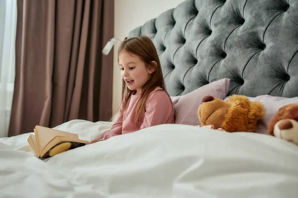 Маленька дівчинка сидить у ліжку в піжамах, покритих ковдрою, читає її книгу і посміхається з плюшевим ведмедем біля неї у величезній світлій спальні вдома — стокове фото