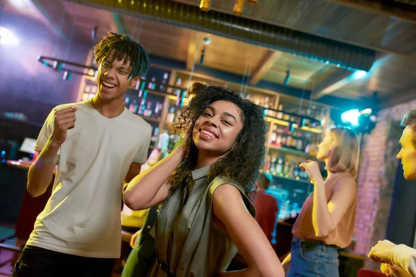 Disfruta de la noche. Atractiva mujer joven de raza mixta posando en la cámara mientras se divierten, bailando con sus amigos en la fiesta en el bar — Foto de Stock