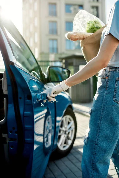 Close-up van de hand van de vrouw in beschermende handschoen openen van de blauwe auto deur terwijl het houden van boodschappenzak, Beschermende maatregelen tegen bacteriën, virussen, coronavirus pandemie — Stockfoto