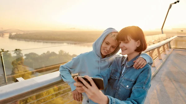 Lesbický pár stojící na mostě, usmívající se při focení selfie, sledující společně východ slunce — Stock fotografie