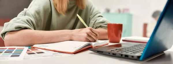 A cute young well-dressed blondy making notes with a pencil at a table in front of a laptop — Stock Photo, Image