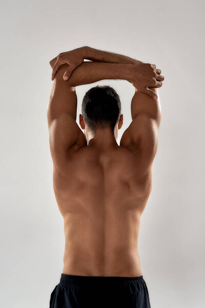 Идеальное мужское тело. Вид сзади на мускулистого человека с обнаженным туловищем, поднимающим руки над головой изолированным на сером фоне, позирующего в студии