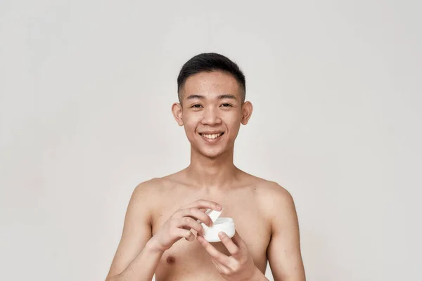 Portrét mladého Asiata s problematickou pletí a hyperpigmentací na tváři, usmívajícího se do kamery, otvírajícího smetanovou sklenici izolovanou na bílém pozadí. Krása, péče o pleť, koncepce léčby — Stock fotografie