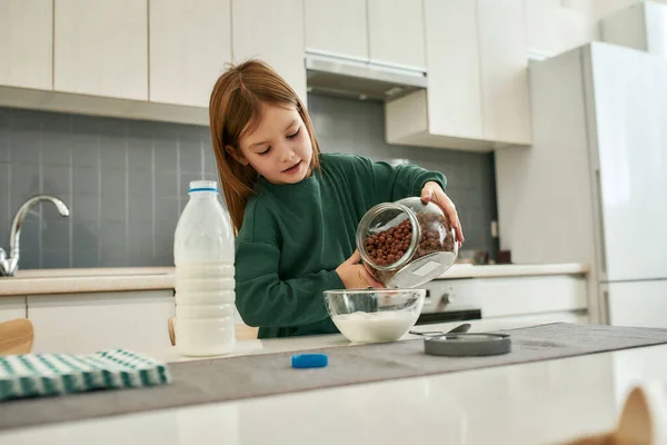 Ein kleines süßes Mädchen gießt in einer großen Küche Müslibällchen in eine Schüssel mit Milchflecken — Stockfoto