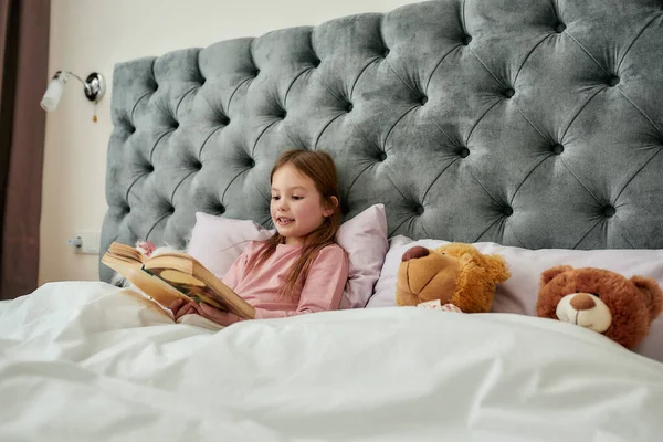 Симпатична маленька дівчинка наполовину лежить на подушках у величезному ліжку, покритому ковдрою, читаючи її книгу вголос — стокове фото
