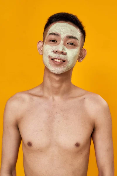Portrait of shirtless young asian man with green mask applied on his face smiling at camera isolated over yellow background. Beauty, skincare routine concept