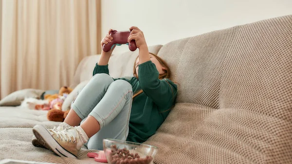 Uma menina segurando um gamepad enquanto joga jogos em um console de TV sentado em um sofá sozinho — Fotografia de Stock