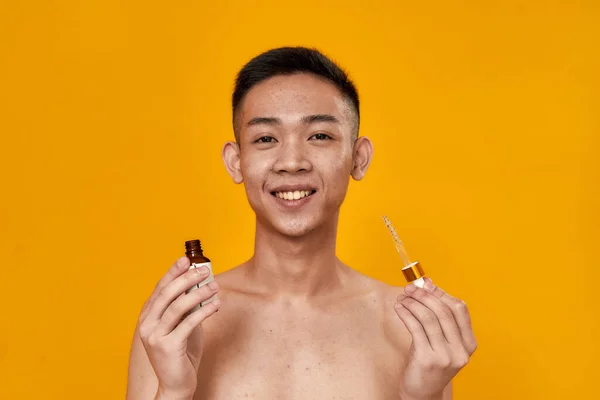Porträtt av ung asiatisk man ler mot kameran medan du håller gel serumflaska och glas pipett isolerad över gul bakgrund — Stockfoto