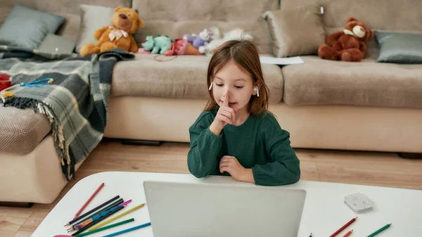 Uma pequena garota bonita mostrando para manter o silêncio enquanto ela está fazendo sua tarefa doméstica sozinha em um laptop sentado a uma mesa sozinha durante a educação à distância — Fotografia de Stock