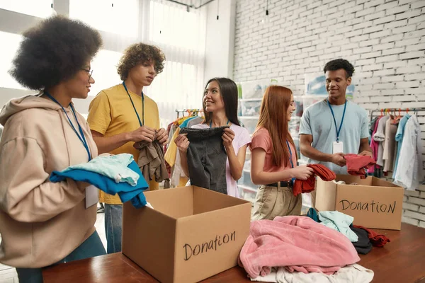 Gelukkig jonge vrijwilligers groep sorteren, verpakken kleding in kartonnen dozen, divers team werken samen aan donatie project in charitatieve organisatie kantoor — Stockfoto