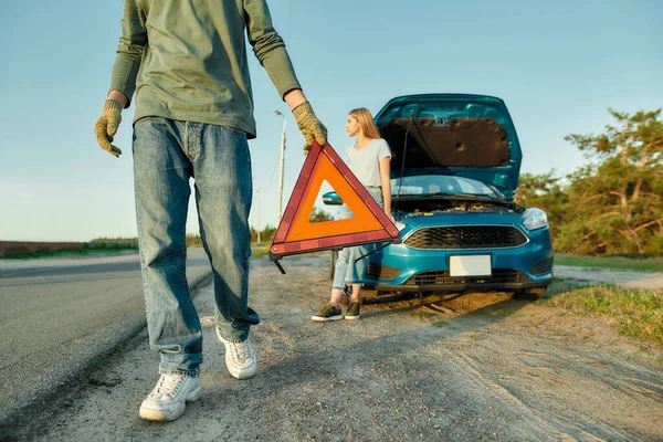 Gehakt schot van de man bestuurder zetten rode waarschuwing driehoek of noodstop teken achter zijn kapotte auto aan de kant van de weg, zijn vriendin wachten op de achtergrond — Stockfoto