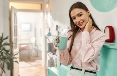 Tasarımcı kıyafeti giymiş, elinde bir fincan çayla kameraya bakıp gülümseyen sevimli, uzun saçlı bir esmer.