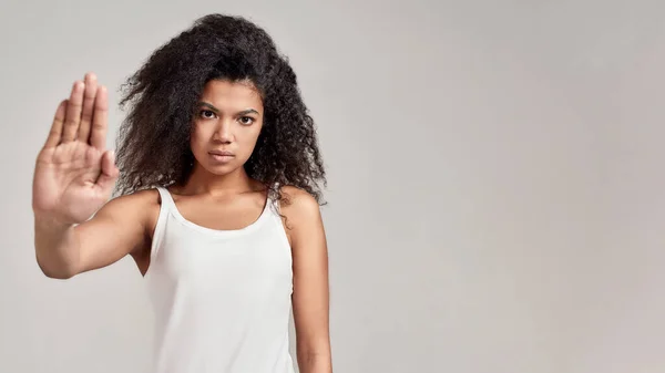 Retrato de jovem afro-americana com cabelo encaracolado vestindo camisa branca com sério olhar confiante ao mostrar, fazendo parar gesto isolado sobre fundo cinza — Fotografia de Stock