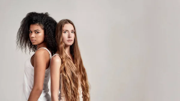 Portret dwóch młodych, zróżnicowanych kobiet ubranych w białe koszule patrzących w kamerę stojących razem na szarym tle — Zdjęcie stockowe