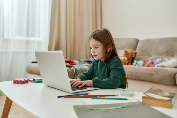 Uma menina pequena passar o tempo sozinha na frente de um laptop com seus brinquedos fofos em um sofá durante a educação à distância — Fotografia de Stock