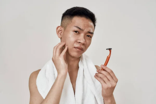 Retrato de un joven asiático con piel problemática sosteniendo una navaja, mirando a la cámara con vacilación aislada sobre fondo blanco. Belleza, cuidado de la piel, rutina de la mañana — Foto de Stock