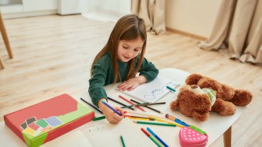 Küçük bir kız masada tek başına oturuyor ve büyük parlak bir odada kalemlerle resim yapıyor. Uzaktan eğitim sırasında gülümsüyor ve resim çizmekten zevk alıyor.