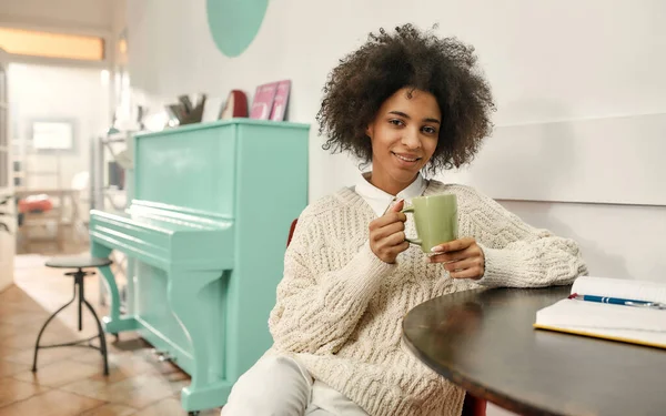 얼굴이 검은 여자가 커피 한 잔을 들고 화려 한 옷을 입고 웃으며 의자에 앉아 있는 카메라를 들여다보고 있습니다 — 스톡 사진