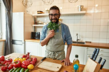 Neşeli genç adam, önlüklü İtalyan aşçı kameraya bakıyor, fesleğen yapraklarını hazır tutuyor sebzelerle sağlıklı bir yemek hazırlıyor, mutfakta duruyor. Ev konseptinde aşçılık