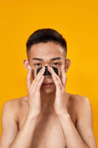 Retrato de un joven asiático sin camisa mirando a la cámara mientras aplica una mascarilla de carbón en su nariz aislada sobre un fondo amarillo. Puntos negros, concepto de tratamiento del acné — Foto de Stock