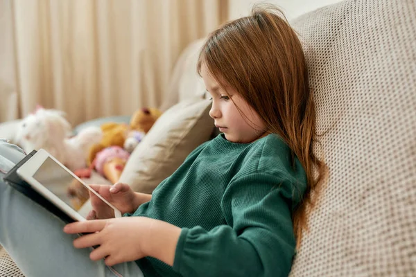 Uma pequena garota fofa totalmente imersa em um videogame em seu tablet enquanto sentada em um sofá — Fotografia de Stock