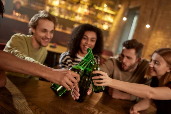 ¡Salud! Un grupo de amigos felices en el bar viendo partidos deportivos en la televisión juntos, bebiendo cerveza, tintineando botellas y animando al equipo. Gente, ocio, amistad y entretenimiento — Foto de Stock