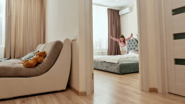 A cute girl jumping down on a big bed in a bedroom having her teddybear in a guestroom — Stock Photo, Image