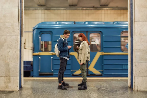 Foto completa de un par de adolescentes usando teléfonos inteligentes, totalmente absortos en la vida en línea, ignorándose mutuamente mientras están de pie en la estación de metro —  Fotos de Stock