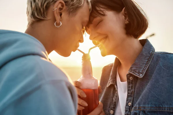 Happy young lesbian couple having romantic moment, drinking from one glass bottle with the straw, Two women enjoying cold beverage on a summer day outdoors — Stock Photo, Image