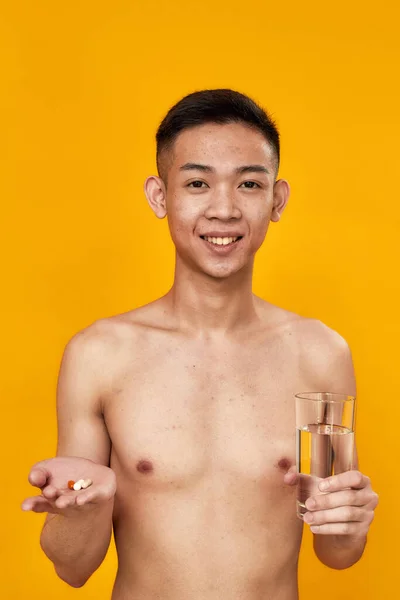 Portret bezkoszulkowego młodego Azjaty uśmiechającego się do aparatu, trzymającego tabletki i szklankę wody na żółtym tle. Opieka zdrowotna, koncepcja leczenia — Zdjęcie stockowe